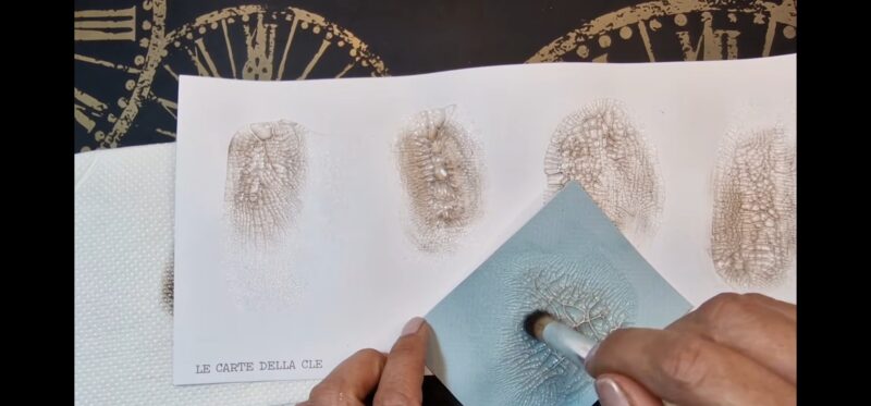 Si vede in primo piano un pennello che passa sopra la carta con l'effetto craklé. Sullo sfondo si vedono altri pezzi di carta con lo stesso effetto