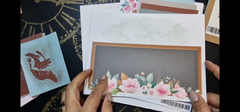 Si vede in primo piano una carta decorata con un riquadro con rettangolare con sfondo nero e bordi marrone. Sul lato inferiore del rettangolo ci sono molti fiori rosa di svariate dimensioni