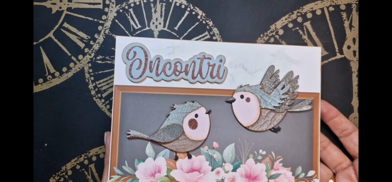 In primo piano si vede il pannello assemblato. Sullo sfondo c'è la carta con la decorazione a finestra e i fiori rosa sulla base, al centro ci sono i due uccellini che si guardano e in alto a sinistra la scritta incontri