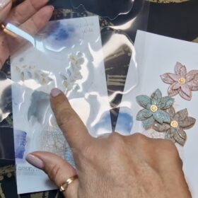 In primo piano si vede un foglio di acetato trasparente e ci sono degli adesivi trasparenti. Sullo sfondo si vede un pannello bianco con il vaso e i fiori craklé
