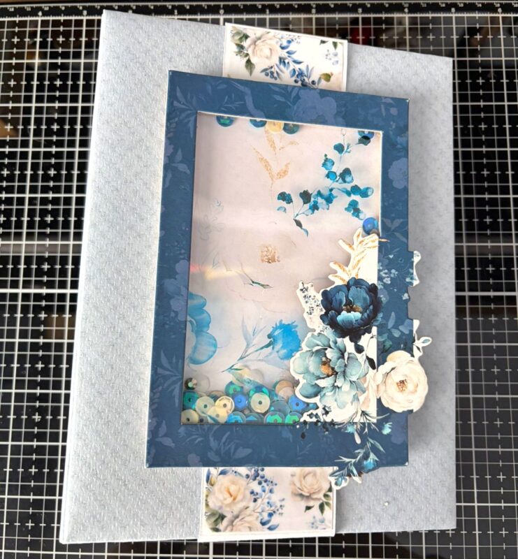 Copertina di un album fotografico fatto a mano con la carta. Colore azzurro, dettaglio di fiori nell'angolo a destra in basso e cornice spessorata con paillettes